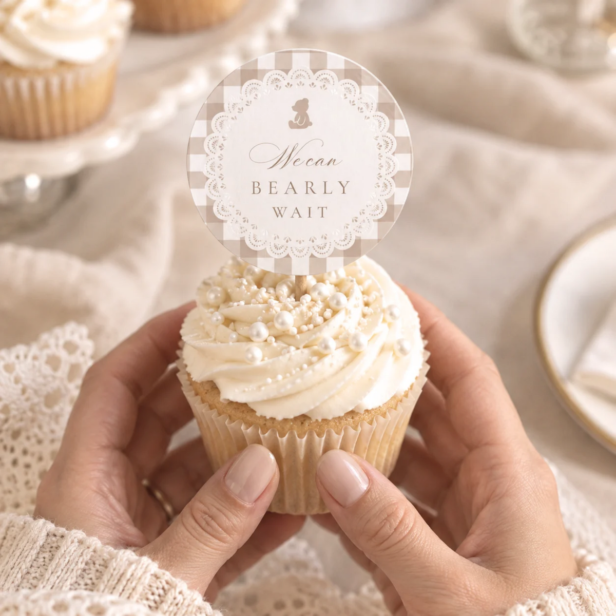 Hands holding a cupcake with a We Can Bearly Wait teddy bear cupcake topper in beige gingham and lace
